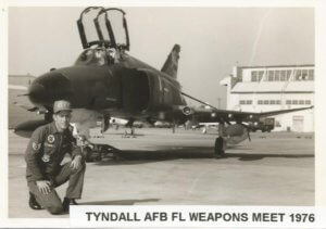 A black and white photo of a man crouching down in front of a warplane on an airfield.