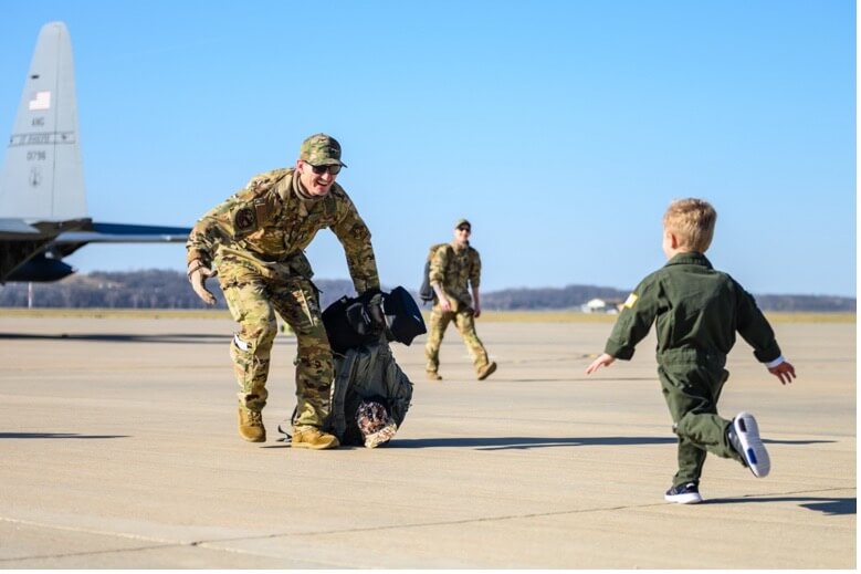 A man in fatigues and sunglasses bending down to greet a young boy running toward him.